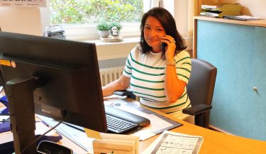Frau Krybus aus dem Sekretariat sitzt an ihrem Schreibtisch und lächelt mit dem Telefonhörer in der Hand.