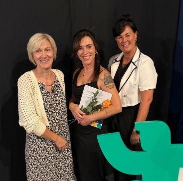 Zu sehen ist Peggy Pinkert mit ihrem Zeugnis, einer Blume und den beiden zentralen Praxisanleiterinnen.