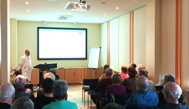 Zu sehen ist ein Blick von hinten nach vorn während der Veranstaltung. Einer der Ärzte hält einen Vortrag vor den Zuhörern.