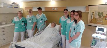 Zu sehen sind die Auszubildenden des 2. Turnus, die im Pflegetrainingszentrum um das Trainingsbett mit Puppe herum stehen.