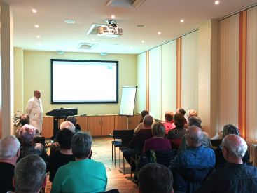 Zu sehen ist ein Blick von hinten nach vorn während der Veranstaltung. Einer der Ärzte hält einen Vortrag vor den Zuhörern.