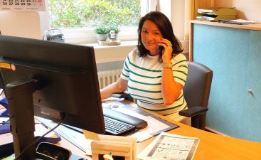Frau Krybus aus dem Sekretariat sitzt an ihrem Schreibtisch und lächelt mit dem Telefonhörer in der Hand.
