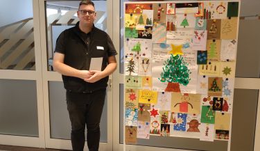 Zu sehen ist der Seelsorger Herr Liebert, der vor der Tafel mit den Weihnachtskarten im Foyer der Klinik steht.