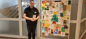 Zu sehen ist der Seelsorger Herr Liebert, der vor der Tafel mit den Weihnachtskarten im Foyer der Klinik steht.